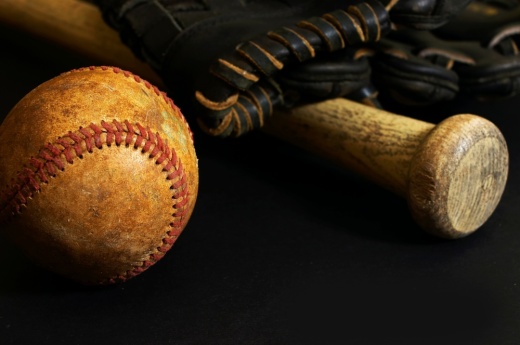 A rustic looking baseball, baseball bat and the edge of a baseball glove are seen in a dimly lit setting.