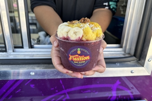 A purple fruit bowl is shown with toppings being handed from inside the food truck to the customer.