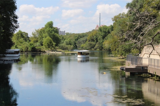 Pictured is a lake with hills covered in trees in the background as well as Texas State University's Old Main building.