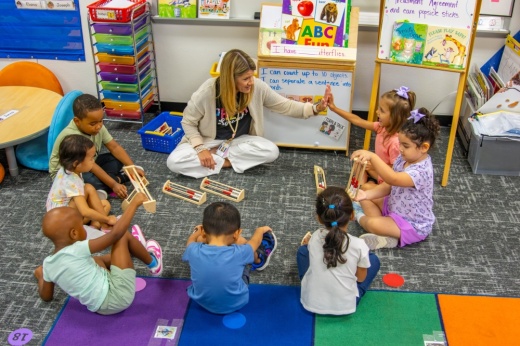 a teacher sits in a circle on the floor with a pre-K class