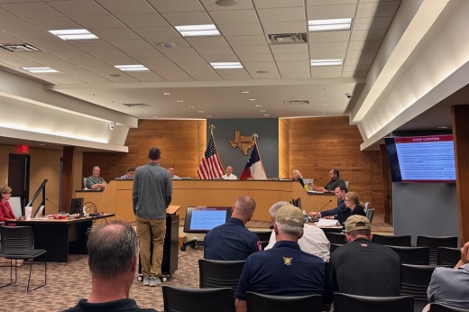 people sit in a city council room and listen to a man speaking at a podium