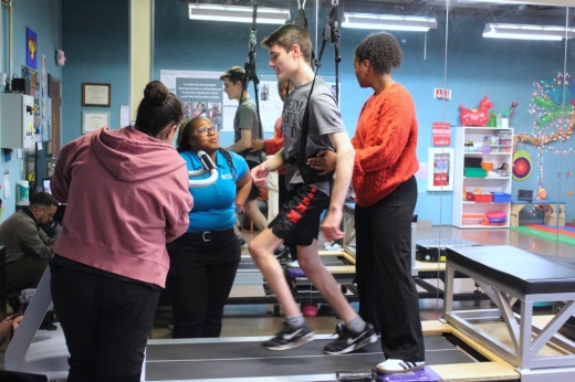 A teenage boy walks on a treadmill in a harness with support from physical therapists