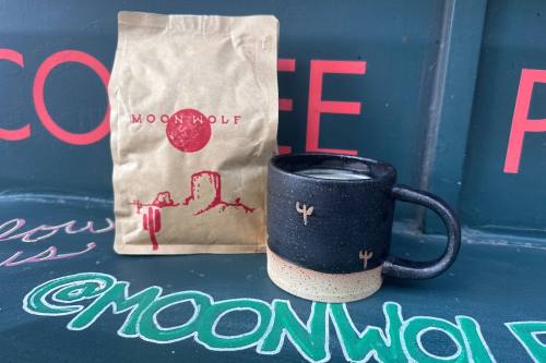 Cup of coffee next to a brown paper bag featuring desert scene graphics and Moon Wolf branding.