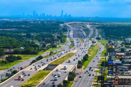 A drone shot shows vehicles along I-35 with a view of Austin's cityscape from Hays County in the background.