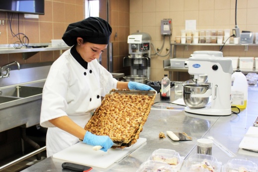 Female wearing a chef's hat and coat while removing a baked item from a tray in an industrial kitchen.