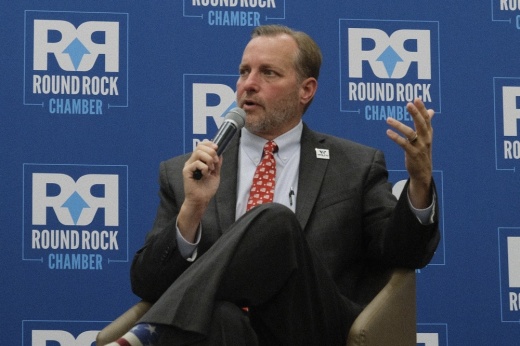 Williamson County Judge Steve Snell holds a microphone while speaking on stage during the State of the County event, seated against a backdrop with the Round Rock Chamber logo.