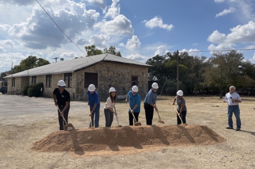 six people wearing white hard hats shovel a pile of dirt