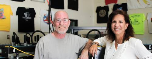 Owner Suzanne Whatley and master screen printer Greg McCoy stand near a screen printing machine.