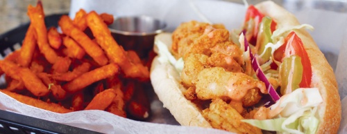 Shrimp pou2019boy, fried shrimp, lettuce, pickles, tomatoes and a spicy remoulade are served on a bun with a side of sweet potato fries. 