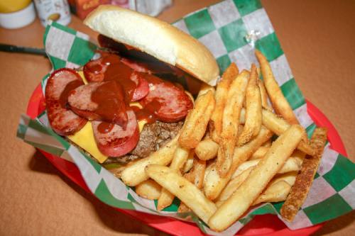 A cheeseburger is seasoned with house spices, served with onion and pickles, and topped with smoked sliced sausage and barbecue sauce for $11.99.